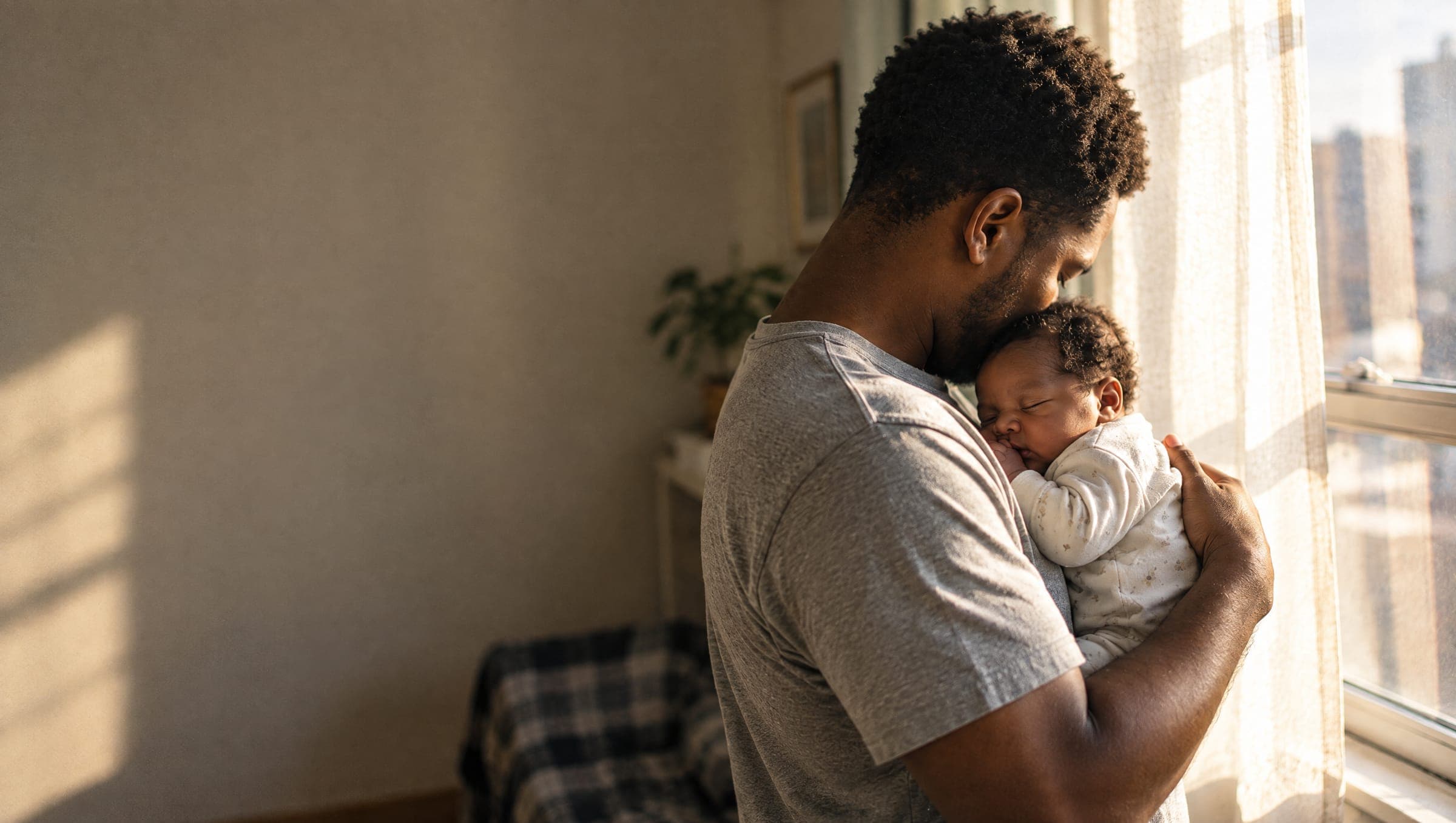 A father holds his sleeping newborn against his chest beside a sunlit window.
